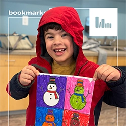 A young child in a red hooded sweatshirt smiles proudly holding up a multi-colored piece of snowman artwork they created