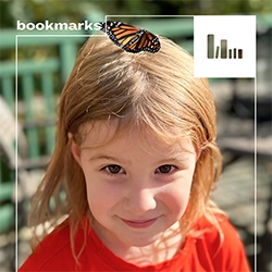 A child looks at the camera with a monarch butterfly in their hair
