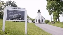 A white church is bookended by a sign for the Church and a large tree